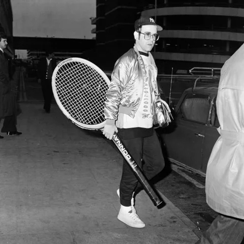 Elton John pictured at Heathrow Airport, arriving back from America, carrying a huge tennis racket, 8th April 1978.