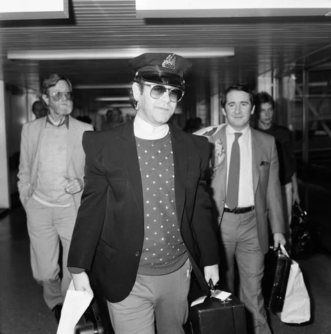 singer elton john arrives at heathrow airport from his 10 week tour of the states wearing an american policemans hat pictured on the right is his manager john reid 21st august 1982