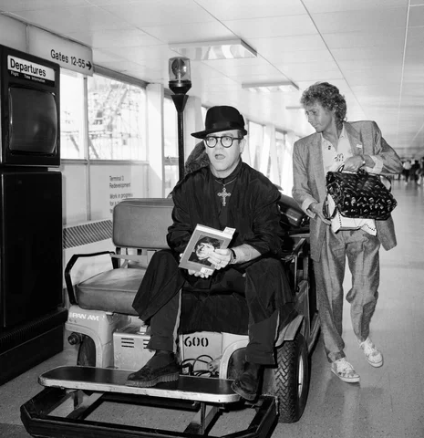 Elton John at Heathrow Airport, wearing a monk's black robe and a big gold cross, topped off with a black hat. He rode on a baggage trolley. He is on his way to California to collect a television award. London. 7th September 1987.